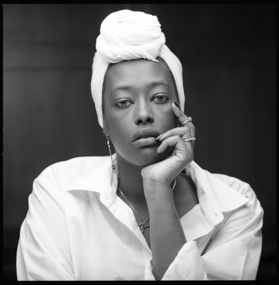 Black and white image of ria resting her face on her hands looking at the camera, wearing a white scarf on her head