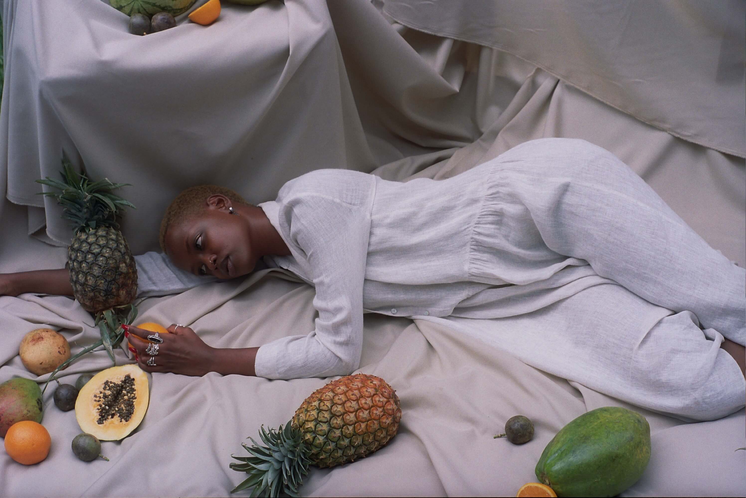 Model lying down on light grey sheets, with fruit scattered around her. She's looking at the pineapple