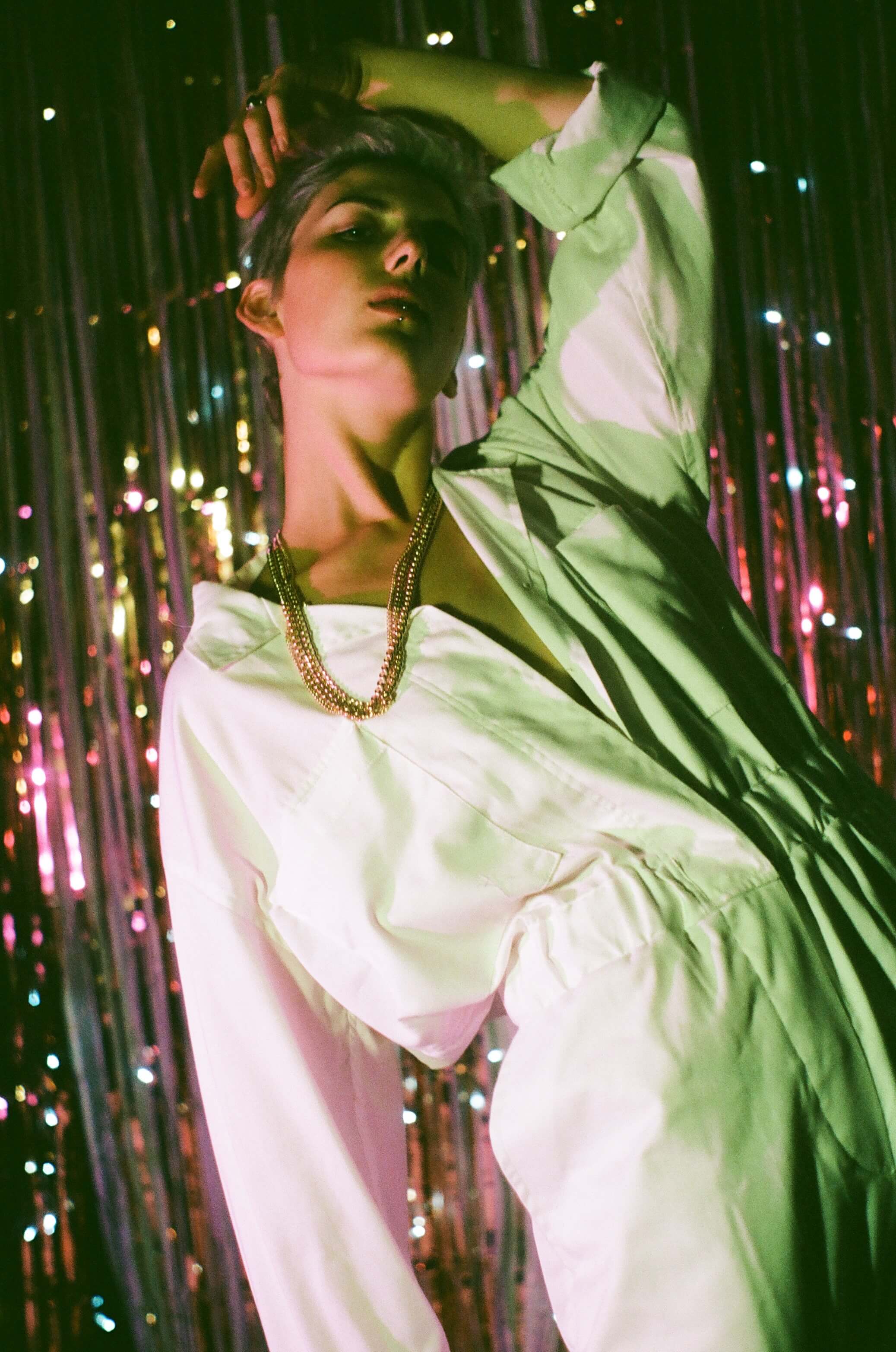 Model facing camera, looking at the camera, standing bent backwards with an arm over their head. Coloured, sparkly background and coloured lighting