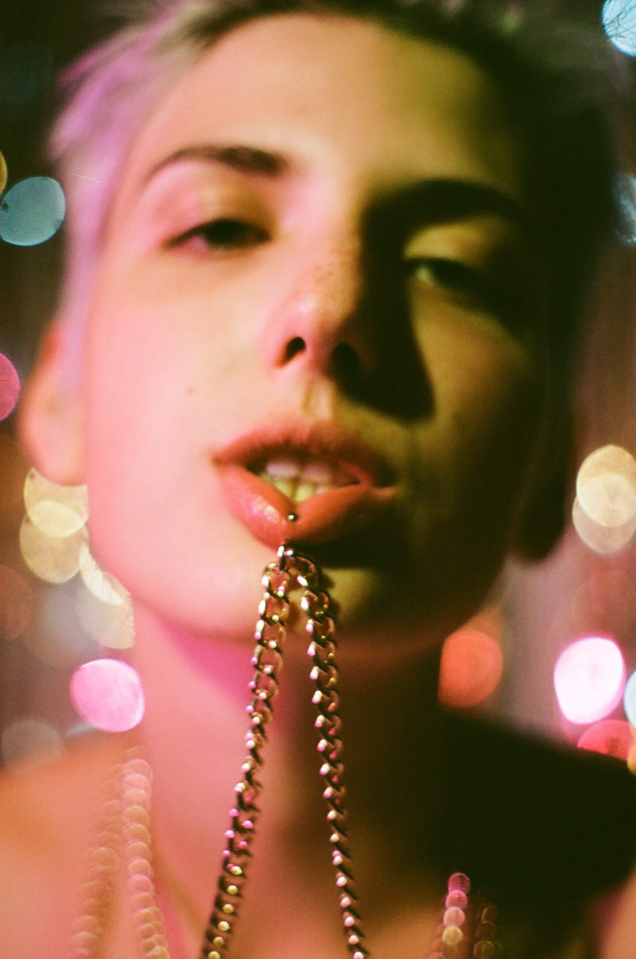 Close up shot of the model's face. A chain attached to their lip piercing, looking straight at the camera. Coloured, sparkly background and coloured lighting