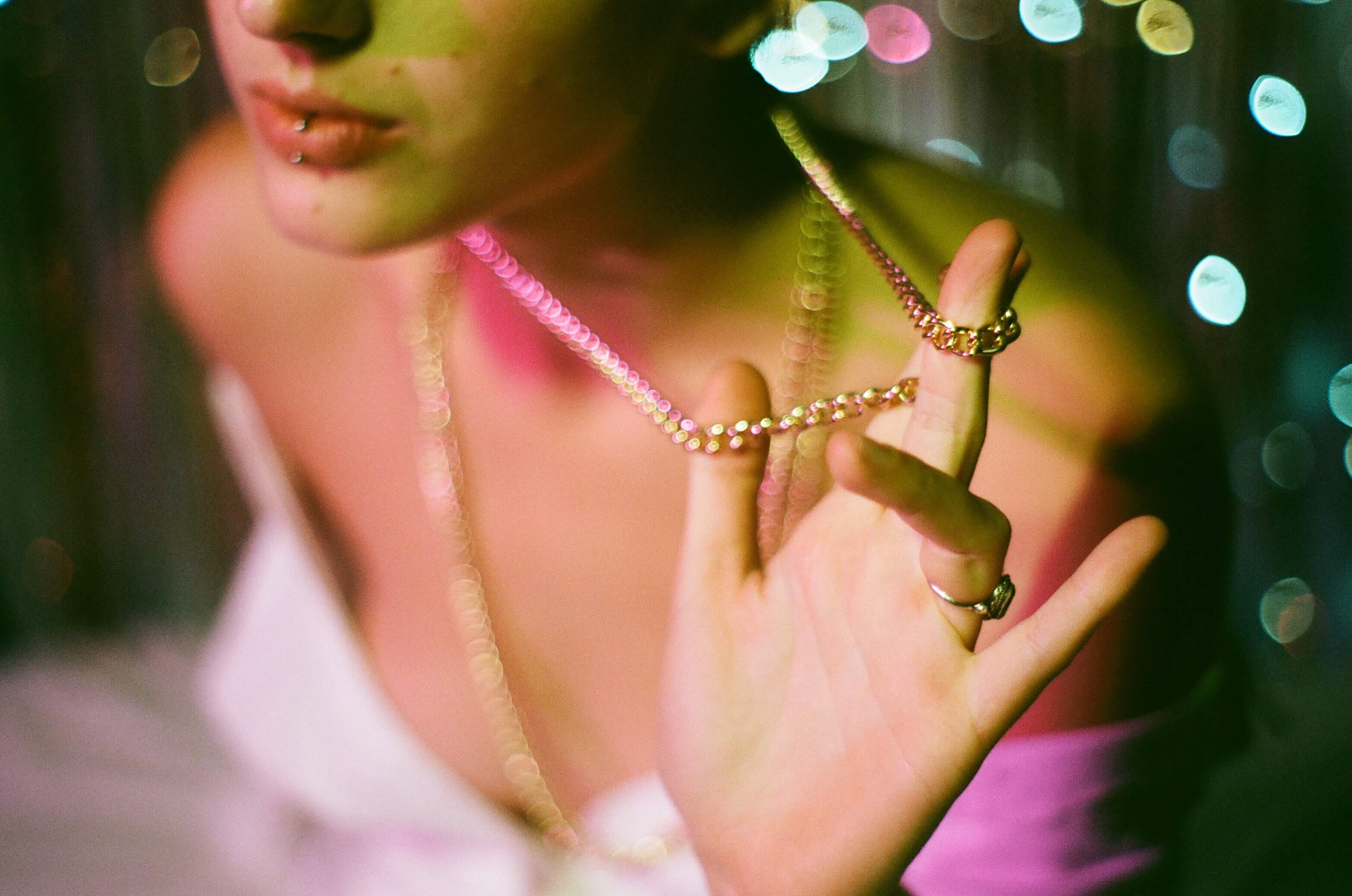 Close up shot of the model's hands playing with their chain necklace. Coloured, sparkly background and coloured lighting