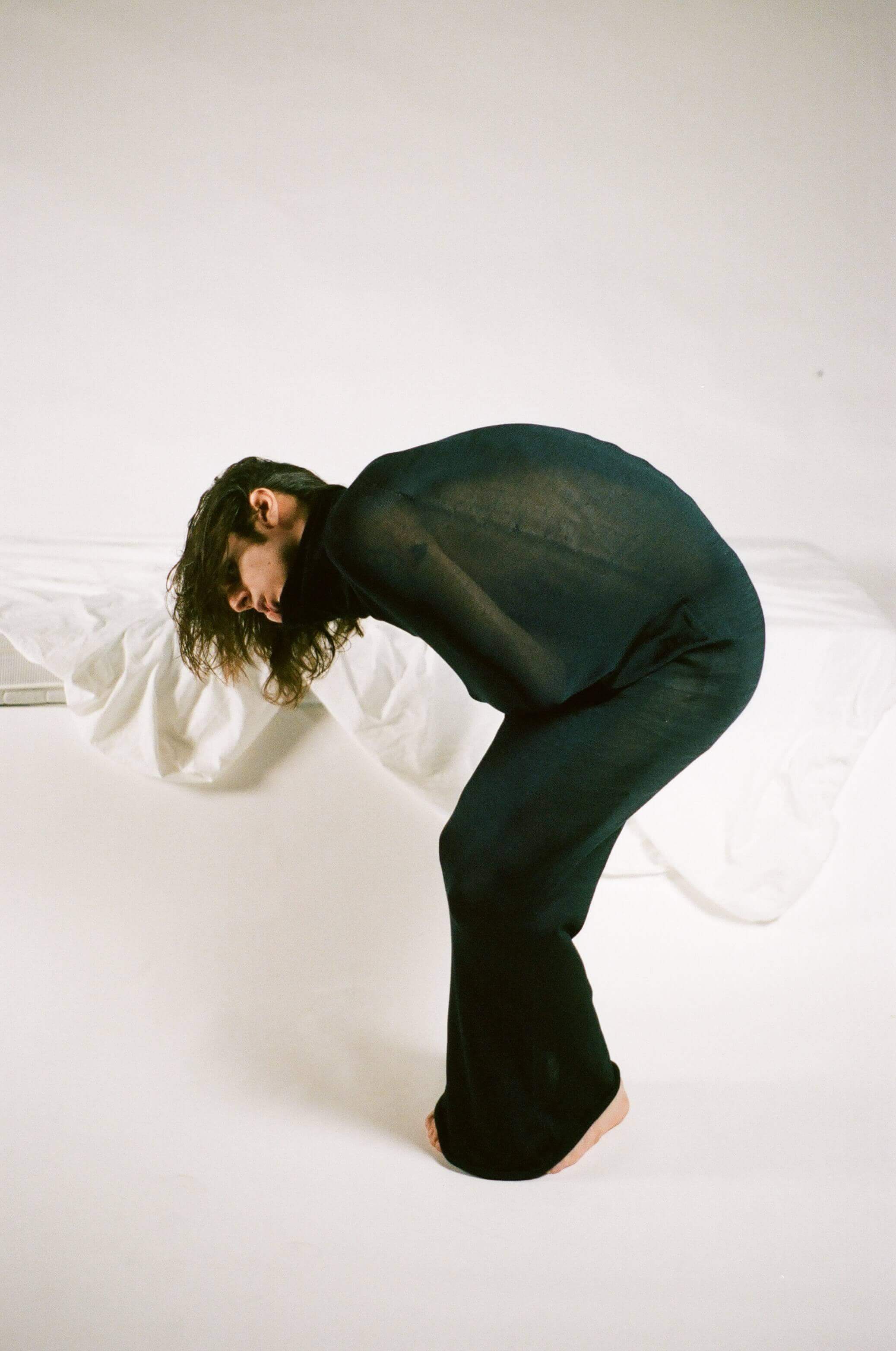 Model wrapped in semi transparent black cloth, bent over in an almost foetal position. White background and bed behind him