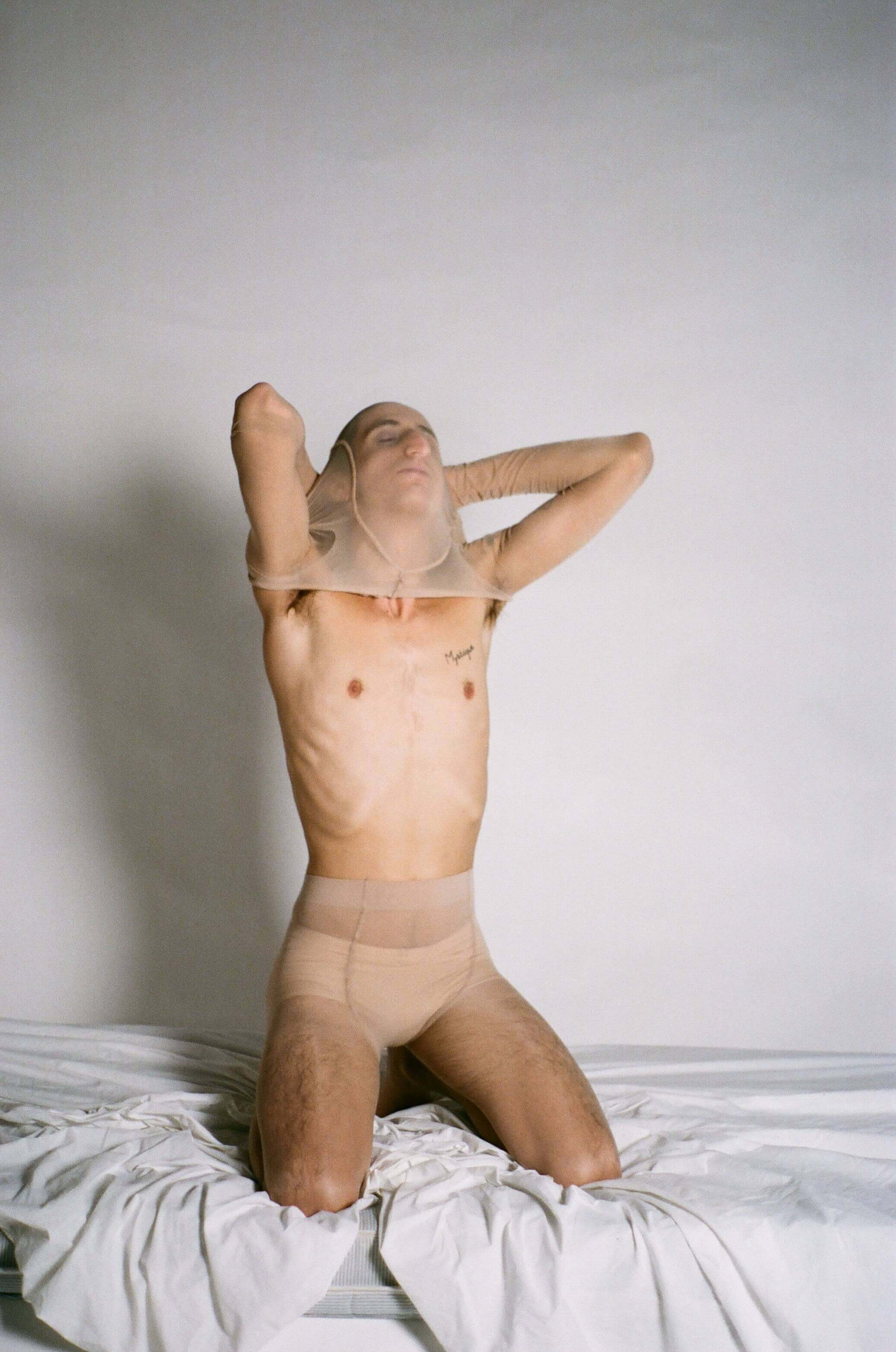 Model wearing beige boxers and tights, kneeling on a bed and holding their head in their hands
